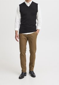 Gilet nero in maglia sopra una camicia bianca con bottoni, abbinato a pantaloni color sabbia e stivaletti in pelle nera. Design a V e dettaglio con bordo a coste.