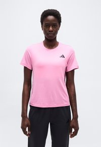 Γυναίκα με ροζ αθλητική μπλούζα με κοντά μανίκια και μαύρο παντελόνι, στέκεται μπροστά σε απλό λευκό φόντο, αντιμετωπίζοντας προς τα εμπρός.