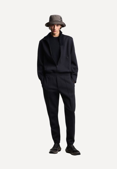 Junger Mann, der eine dunkle Zip-Jacke, passende Hose, schwarze Sneakers und einen grauen Bucket-Hat trägt, steht mit den Händen in den Taschen.