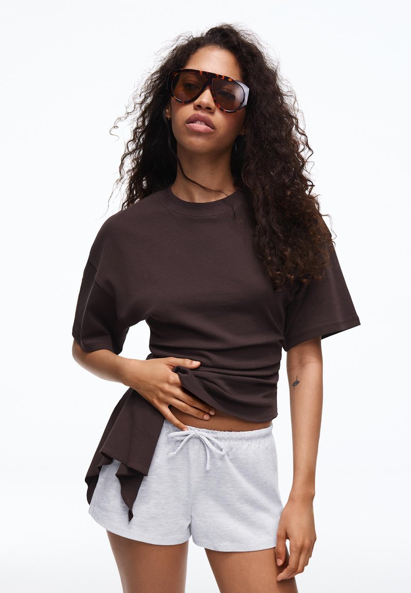 Femme aux longs cheveux bouclés portant de grandes lunettes de soleil écaille de tortue, un t-shirt sombre oversize et un short gris clair à cordon, ajustant son t-shirt.