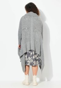 Grå ribbad cardigan med en draperad silhuett, hög krage och utskärningsdetaljer. Buren över en blommönstrad grå klänning och beige stövlar.