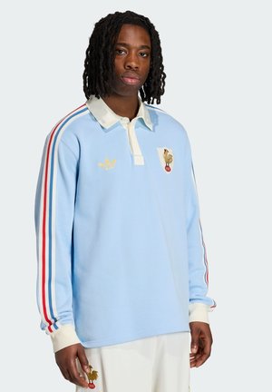 Mladý muž se středně dlouhými copy, nosící světle modré polokošili s dlouhým rukávem se červenými, bílými a modrými pruhy a zlatými logy kohouta.