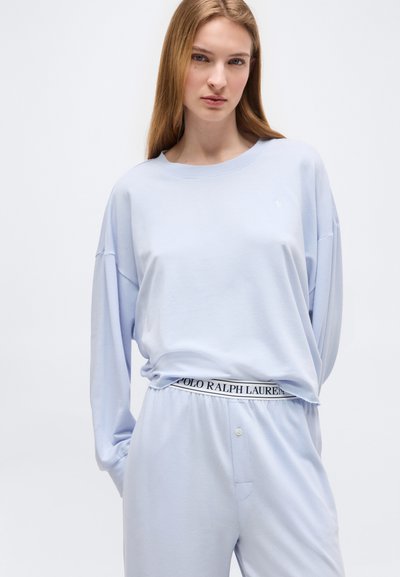 Γυναίκα που φοράει ανοιχτό μπλε μπλουζάκι με μακριά μανίκια και ταιριαστό παντελόνι με ορατό λογότυπο Polo Ralph Lauren στη ζώνη, στέκεται μπροστά σε απλό φόντο.