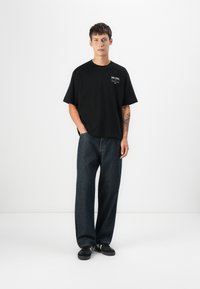 Sort bomulds t-shirt med korte ærmer og løs pasform, parret med mørke denim jeans. Modellen har sorte sneakers på og står foran en hvid baggrund.