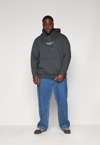 Sudadera gris oscuro con un bolsillo tipo canguro, capucha ajustable con cordón y texto impreso en blanco en el pecho, combinada con jeans azules holgados.