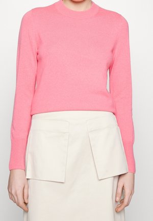 Woman wearing a bright pink long-sleeve sweater and a light beige skirt with two large front pockets, shown from neck to mid-thigh.