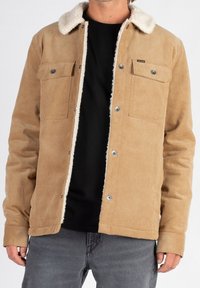 Kurtka z kordurowej tkaniny w odcieniu brązowym z kołnierzem z owczej wełny, z napami, dwoma kieszeniami na piersi i długimi rękawami. Nosi się ją na czarnej koszuli.