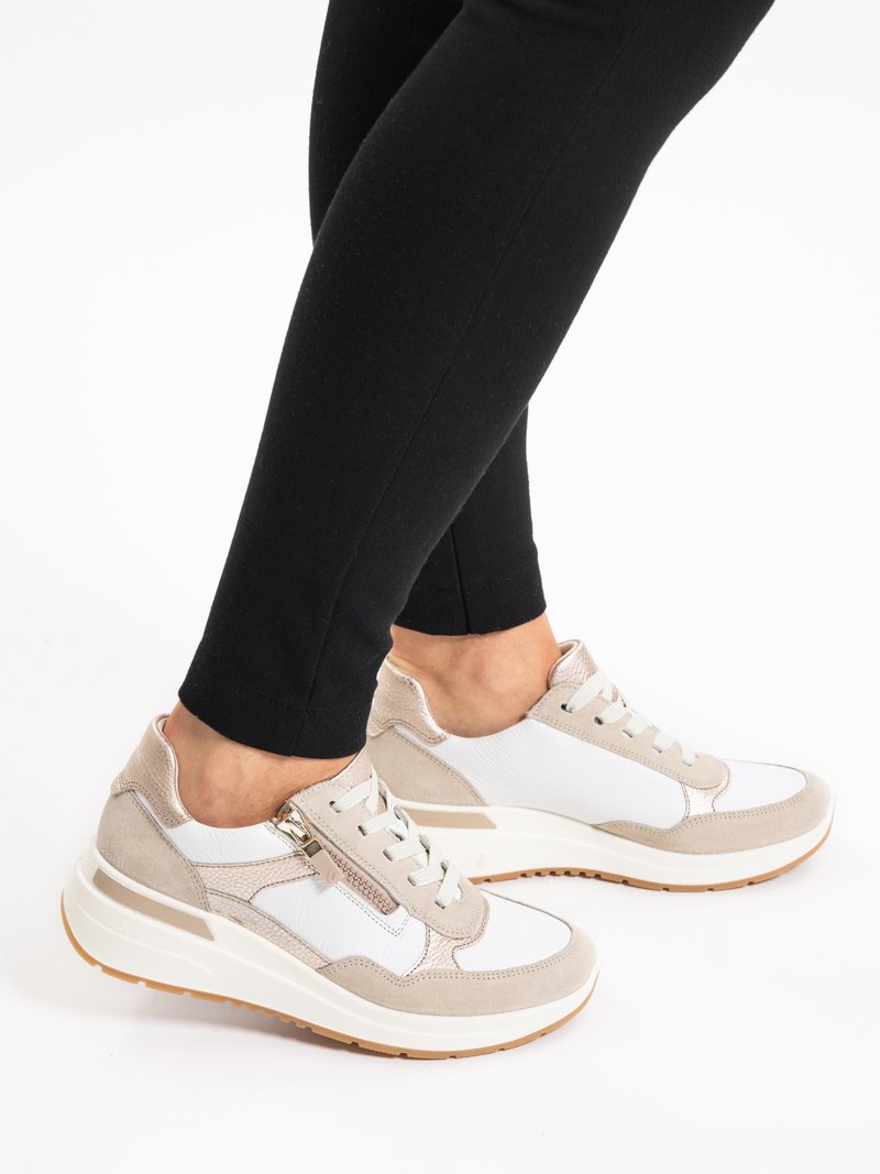 Voeten met witte en beige vetersneakers met zijritsen en gumzolen, gecombineerd met zwarte leggings tegen een witte achtergrond.