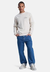 Sudadera beige con cuello redondo, mangas largas y detalle del logo en el pecho, combinada con unos vaqueros azules de corte holgado y zapatillas deportivas grises.