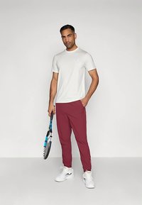 Homem a usar uma t-shirt branca, calças de jogging burgundy e sapatilhas brancas, segurando uma raquete de ténis, de pé contra um fundo cinza claro liso.