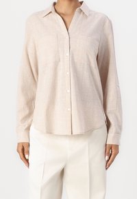 Chemise beige à boutons avec manches longues, un col, deux poches frontales et un motif texturé. Fabriquée en tissu léger.