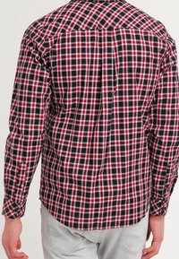 Mies, who wears a longsleeve red, black, and white plaid shirt paired with light gray pants, shown from the back.