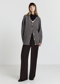 Grau gestrickte Strickjacke mit V-Ausschnitt und sechs großen Knöpfen, kombiniert mit schwarzen Hosen mit weitem Bein, vor hellem Hintergrund.