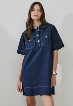 Kvinde iført en kortærmet marineblå denimkjole med knaplukning foran, brystlommer og et armbåndsur på venstre håndled.