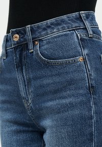 Κοντινή λήψη μπλε τζιν τζιν με μπροστινή τσέπη, θηλιές ζώνης και μεταλλικό κουμπί, φορεμένα με μαύρο τοπ.