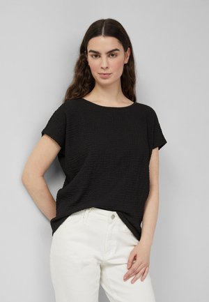 Kobieta o długich ciemnych włosach, ubrana w czarną, teksturowaną bluzkę z krótkim rękawem oraz białe spodnie, stojąca na tle jednolitego szarego tła.