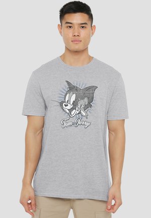 Junger Mann trägt ein hellgraues T-Shirt mit schwarz-weißen Tom-und-Jerry-Cartoon-Gesichtern und Text auf der Vorderseite, steht vor einem schlichten Hintergrund.