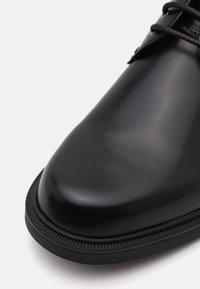 Scarpa di pelle nera con una finitura liscia e lucida, punta arrotondata e una suola in gomma nera testurizzata con un dettaglio a bordo rialzato.