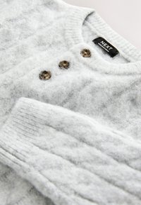 Pull gris en maille fabriqué en matériau doux avec une finition texturée, doté de trois boutons marron et de poignets côtelés. Étiquette de marque visible.