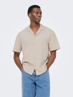 Homme les mains dans les poches portant une chemise beige à manches courtes et un jean bleu, regardant sur le côté devant un fond uni.