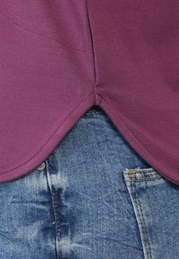 Lila Baumwolloberteil mit abgerundetem Saum, über einer ausgebleichten Jeans mit Distressed-Effekten und gelben Nähten an den Taschen.