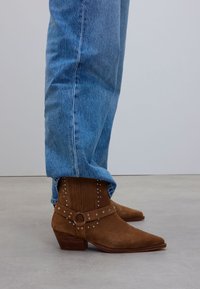Brown suede cowboy boots with pointed toes, featuring a studded strap and textured shaft, paired with light blue denim jeans.