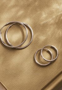 Zilveren oorbellen in twee maten op een gestructureerde, gouden stof achtergrond. De grotere paar is rond, en de kleinere heeft een iets kleinere diameter.