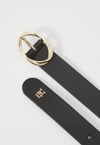 Zwarte leren riem met een gouden ovale gesp. Heeft een gladde textuur en een reliëflogo op de band. Eenvoudig en modern ontwerp.