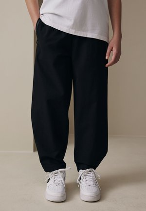 Persona vistiendo pantalones negros holgados, zapatillas blancas con un logo negro, y una camiseta blanca, de pie sobre un suelo liso beige.