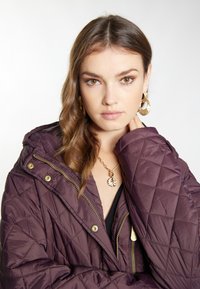 Prošívaná, tmavě fialová péřovka se zlatými doplňky a kapucí. Modelka má na sobě zlaté kruhové náušnice a řetízek s náhrdelníkem ve tvaru mušle.