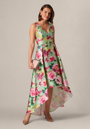 Donna in un abito senza maniche verde menta con stampa floreale, con orlo asimmetrico, che tiene una pochette beige, indossa tacchi beige, in piedi davanti a uno sfondo semplice.