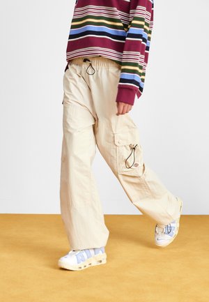 Persona che cammina indossando pantaloni cargo beige, sneakers bianche e blu, e una maglietta a righe burgundy con maniche lunghe su un pavimento senape.