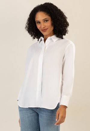 Frau mit lockigem Haar, die ein weißes Hemd mit Knöpfen und blaue Jeans trägt, leicht lächelnd, vor einem einfarbigen beigen Hintergrund.