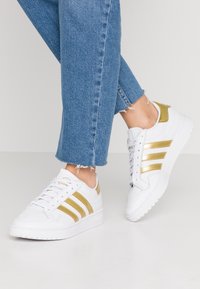 Nohy v modrých zkrácených džínách, obuté v bílých teniskách s metalickými zlatými pruhy a detaily na světlém pozadí.