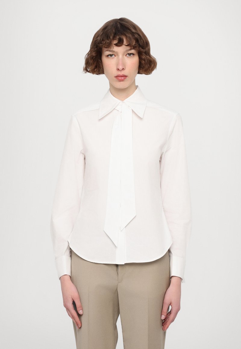 Cameriera bianca a maniche lunghe con colletto e un nastro annodato come accento, caratterizzata da una texture di tessuto liscia e un orlo curvo. Abbinata a pantaloni beige.