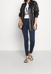 Mujer vestida con una chaqueta de cuero sintético negra, camiseta gráfica blanca, jeans ajustados oscuros, tacones negros con tiras y sosteniendo un bolso de cadena beige.