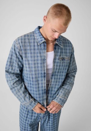 Jeune homme portant une veste à carreaux bleue et un pantalon assorti regarde vers le bas en fermant la fermeture éclair de la veste, sur un fond clair uni.