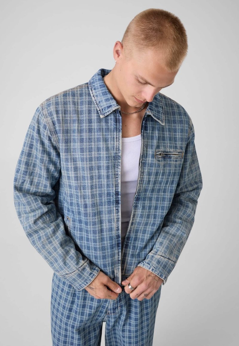 Jeune homme portant une veste à carreaux bleue et un pantalon assorti regarde vers le bas en fermant la fermeture éclair de la veste, sur un fond clair uni.