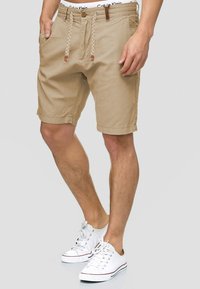 Beige shorts with a relaxed fit, drawstring closure, side pockets, and contrasting stitching. Worn with white sneakers.