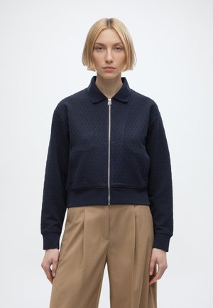 Femme blonde portant une veste matelassée bleu marine zippée avec col et un pantalon large beige, debout devant un fond blanc uni.