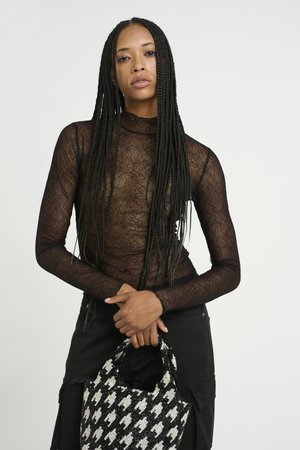Haut noir en dentelle à manches longues avec une texture transparente. Le modèle tient un sac à main en houndstooth noir et blanc orné de perles, assorti à un pantalon sombre.