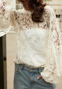 Blonde blonde i creme med lange, lette ærmer og et blomstermønster, lagt over en glat hvid stof. Kombineret med højvaskede denimjeans.