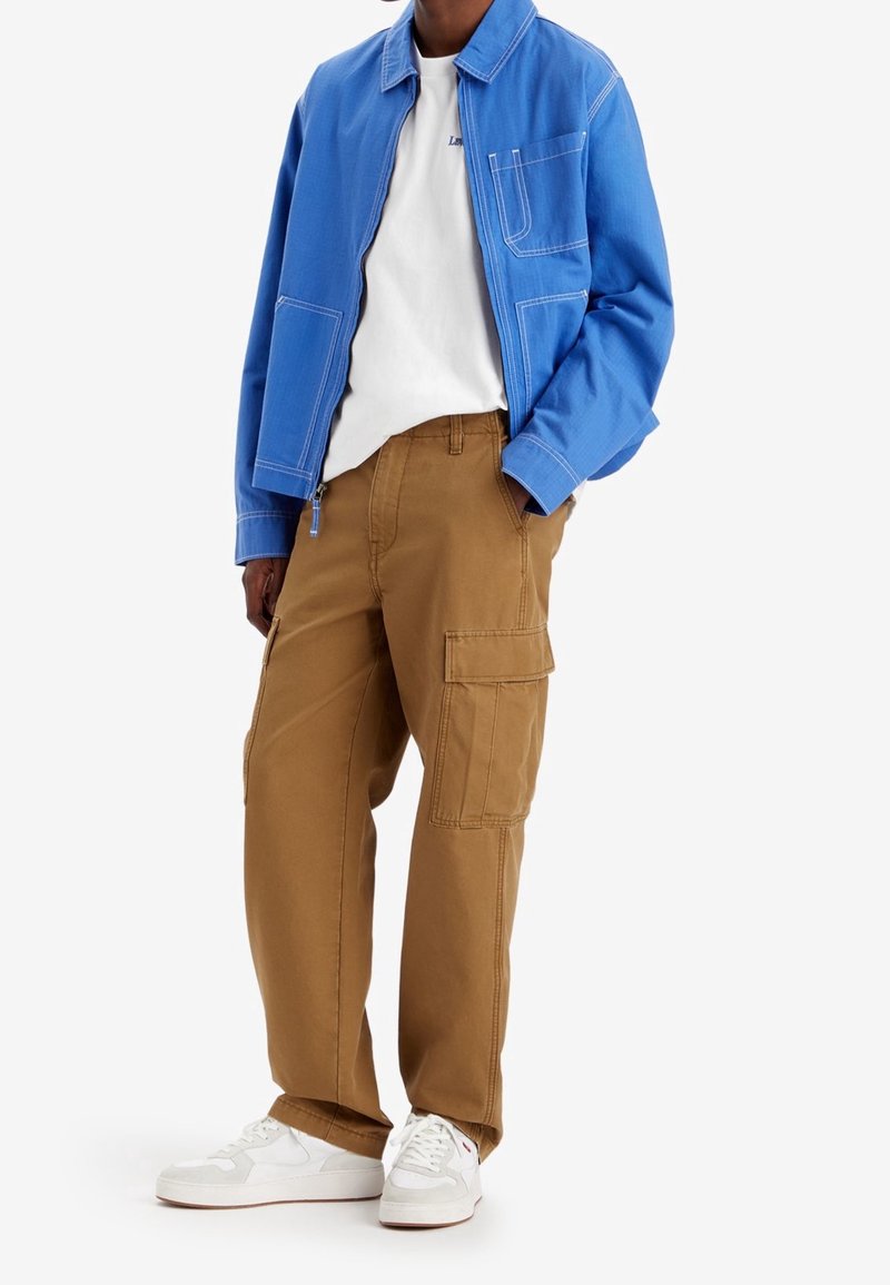 Blå zip-up jacka med vit sömnad, vit t-shirt och beige cargobyxor med sidofickor, kombinerat med vita sneakers.