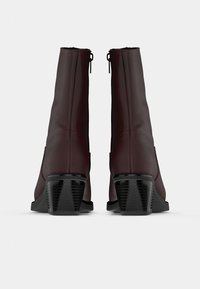 Par de botas de tobillo marrón oscuro con tacones cuadrados negros, mostradas desde la parte trasera sobre un fondo blanco.