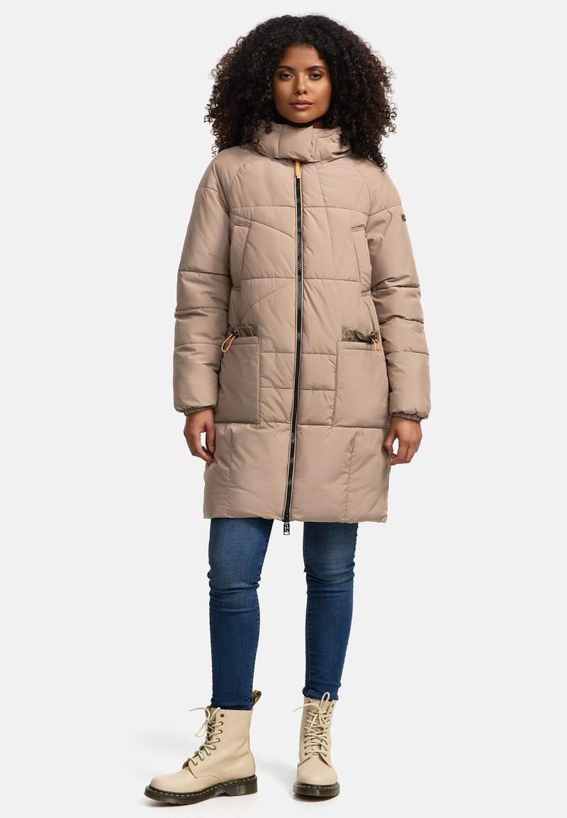 Beige gewatteerde jas met een hoge kraag, vooraan met een ritsluiting, twee voorzakken en toggles; gecombineerd met blauwe jeans en beige laarzen.