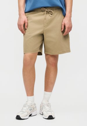 Persoon draagt beige shorts met trekkoord, witte sokken en witte New Balance sneakers, staand voor een effen achtergrond.