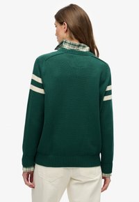 Zielony dziany sweter z białymi paskami na rękawach, teksturowaną tkaniną i częściowo widocznym kołnierzykiem koszuli w kratę pod spodem.