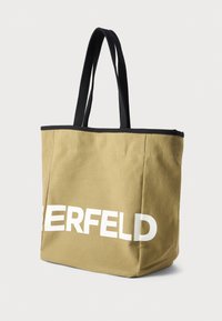 Geantă din pânză bej cu mânere și margini negre. Text mare alb pe lateral. Formă rectangulară, material rezistent și design minimalist.