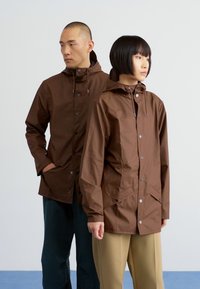 Des vestes imperméables marron avec des capuchons, des fermetures à bouton et des poches avant, présentant une texture lisse et une coupe décontractée, portées par deux mannequins.