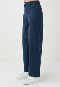 Chabrowe spodnie o szerokich nogawkach o gładkiej teksturze, z pojedynczym guzikiem z przodu i dwiema bocznymi kieszeniami; zestawione z białymi trampkami.